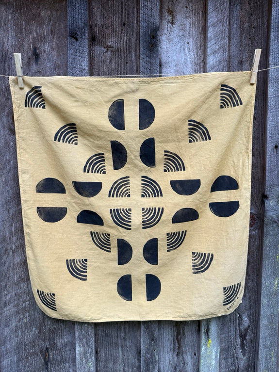 A yellow organic cotton bandana with black block prints, hanging on a wooden fence.